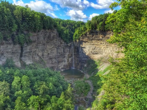 A scenic view of a waterfall cascading into a lush green gorge surrounded by trees and rocky cliffs under a blue sky.