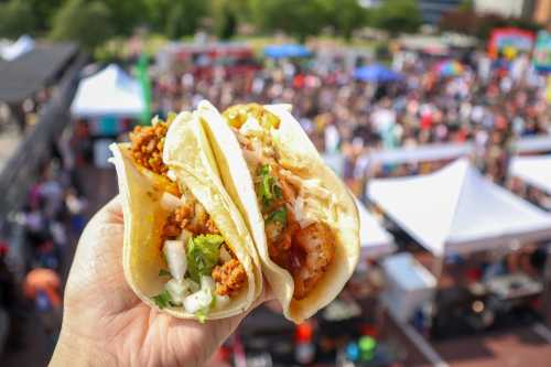 A hand holding three tacos with various fillings, set against a blurred crowd at a food festival.