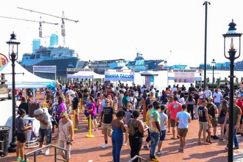 A bustling outdoor event with crowds enjoying food stalls by the waterfront, featuring a large ship in the background.