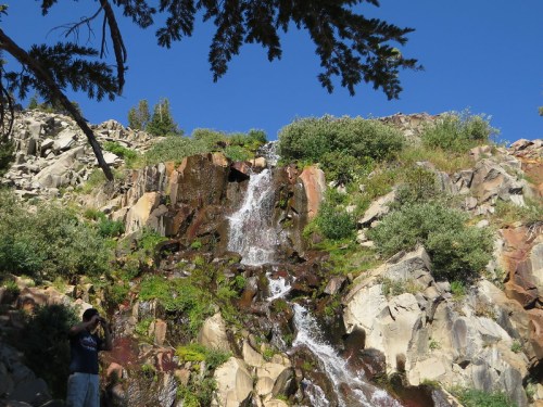 A cascading waterfall flows down rocky terrain, surrounded by lush greenery and a clear blue sky.