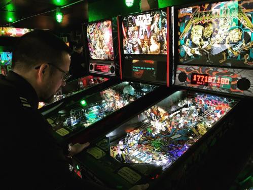 A person playing a pinball machine in a vibrant arcade filled with colorful game displays.