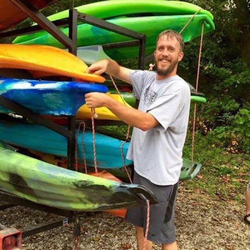 A man stands next to a trailer loaded with colorful kayaks, smiling and holding one of the kayaks.