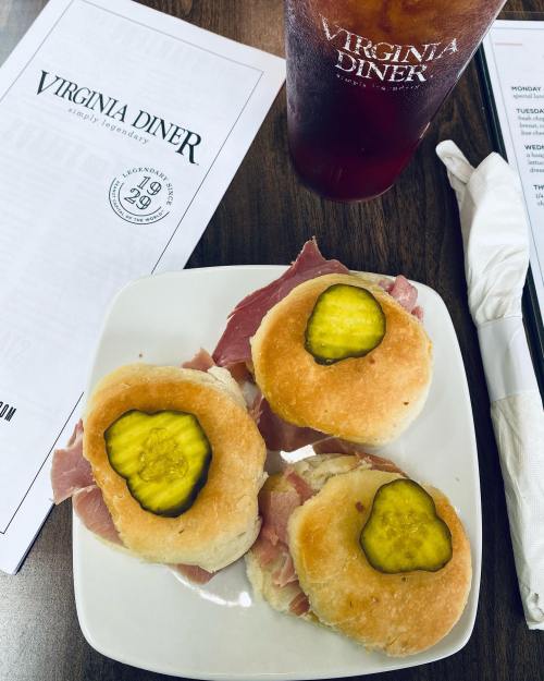 Three small sandwiches with ham and pickles on a white plate, accompanied by a menu and a glass of iced tea.