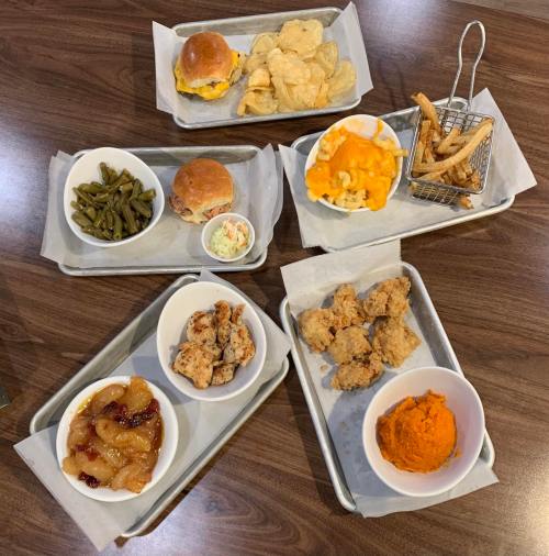 A spread of comfort food including fried chicken, macaroni and cheese, green beans, rolls, and sweet potatoes on metal trays.