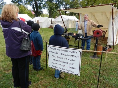 A blacksmith demonstrates metalworking to a small group, with a sign explaining his craft in the background.