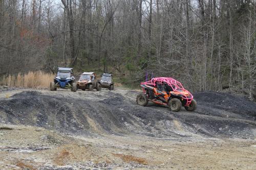 Four off-road vehicles navigate a rugged, muddy terrain surrounded by trees and sparse vegetation.