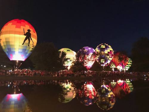 Colorful hot air balloons illuminated at night, reflecting in a calm water surface, with a crowd gathered nearby.