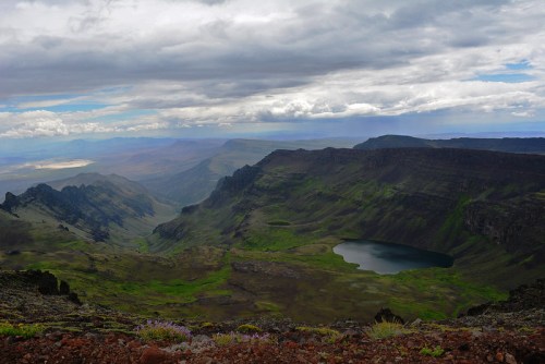 A panoramic view of a mountainous landscape with a lake surrounded by green valleys under a cloudy sky.