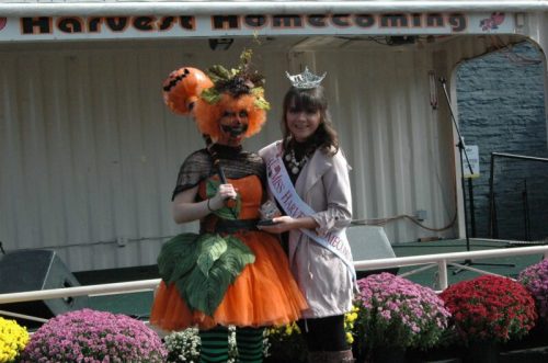 A person in a pumpkin costume poses with a pageant winner at a Harvest Homecoming event, surrounded by flowers.