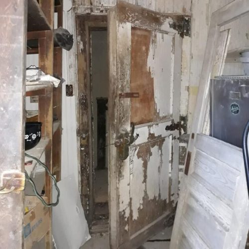 An old, weathered door in a dimly lit room, surrounded by shelves and various objects.