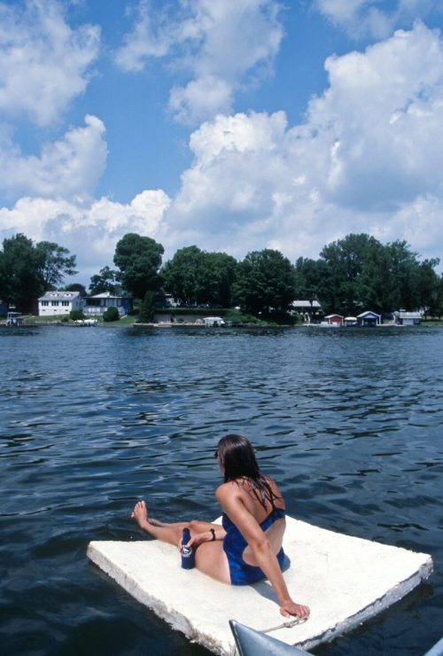 A person sits on a floating platform in a lake, gazing at the shoreline under a partly cloudy sky.
