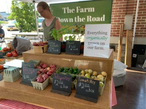 A farmer's market stand displays fresh produce, including shallots, basil, garlic, jalapeños, and tomatillos.