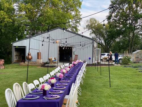 A long table set with purple linens and flowers, outside a barn with string lights, surrounded by greenery.