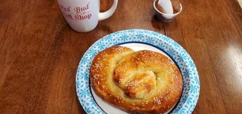 A cinnamon roll on a paper plate, with a coffee mug and a small container of sugar on a wooden table.