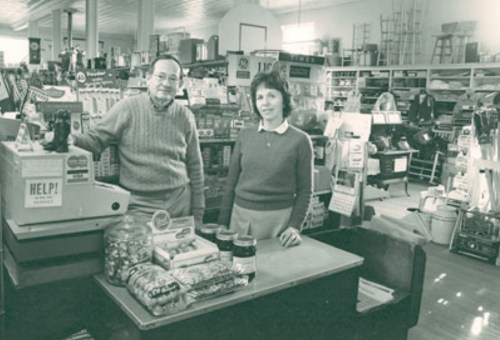 A man and woman stand behind a counter in a vintage store filled with various products and shelves.