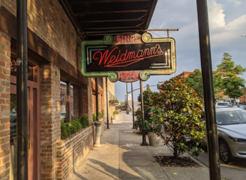 Neon sign for Weldmann's restaurant, with brick building and sidewalk lined with greenery and parked cars.