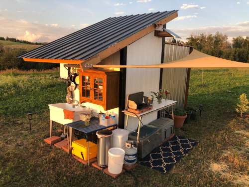 A small, modern cabin with a metal roof, outdoor seating, and a shaded area, set in a grassy field.