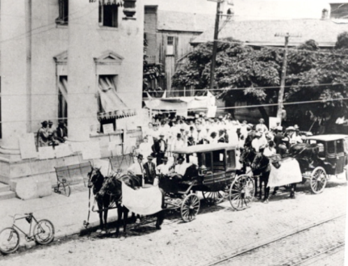 Historic black-and-white photo of horse-drawn carriages and a crowd outside a building, with a bicycle nearby.