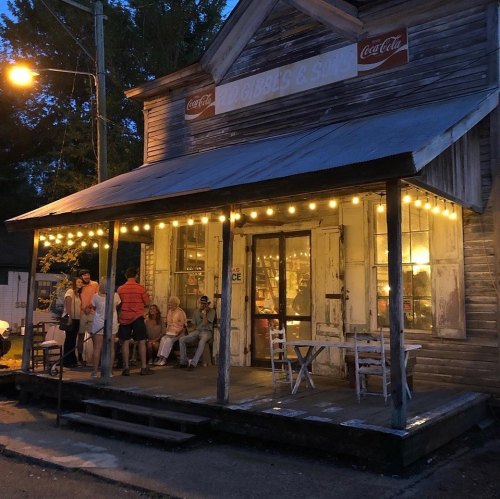 A rustic storefront with string lights, people gathered outside, and a warm glow from the windows at dusk.