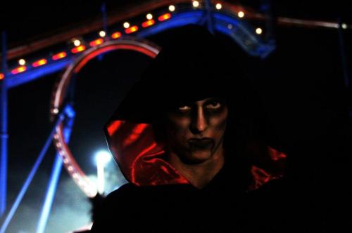 A figure in a dark hooded cloak with dramatic makeup stands against a brightly lit amusement park ride at night.