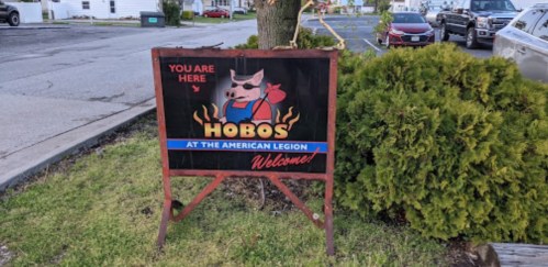 Sign for "Hobos at the American Legion" with a cartoon pig, stating "You Are Here" and welcoming visitors.