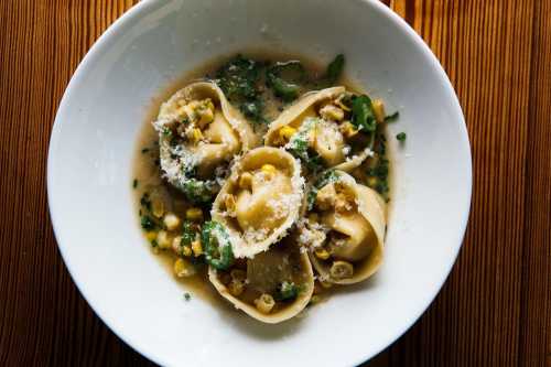 A bowl of pasta filled with corn and greens, topped with grated cheese and served in a light broth.