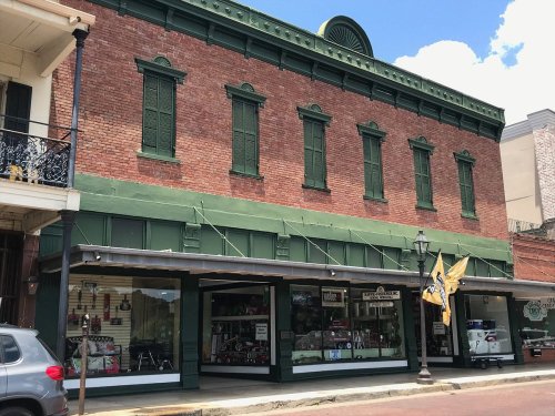 Historic brick building with green shutters, featuring a storefront displaying various items and a parked car outside.