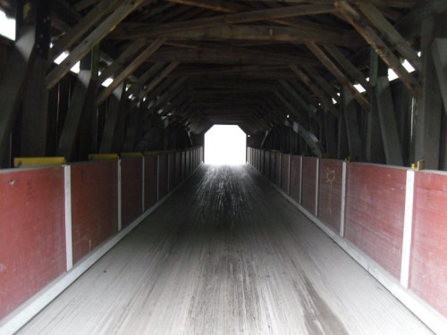 A view down the center of a covered bridge, leading to a bright light at the far end. Wooden beams and red walls visible.