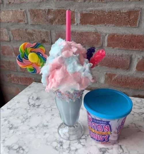 A colorful milkshake topped with cotton candy, lollipops, and sprinkles, next to a container of cotton candy.