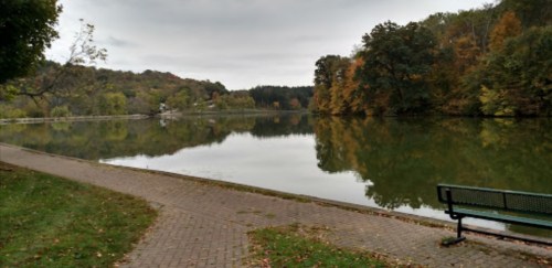 A serene lake surrounded by trees in autumn colors, with a path and a bench along the water's edge.