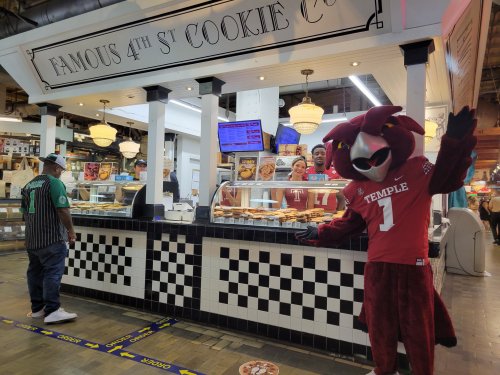 A cookie shop with a mascot in a red jersey waving, while customers browse the display of baked goods.