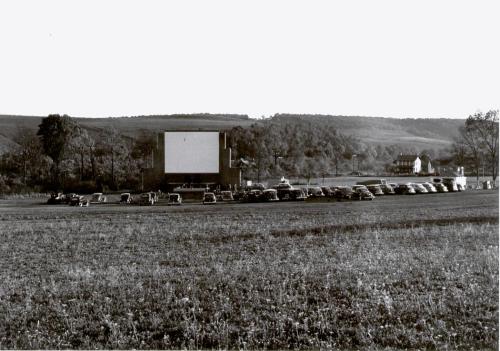 A vintage drive-in theater with parked cars facing a large blank screen, surrounded by fields and hills.