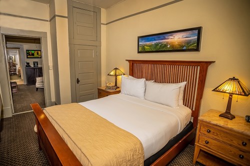 Cozy hotel room with a queen bed, wooden furniture, and a scenic painting on the wall. Doorway leads to another room.