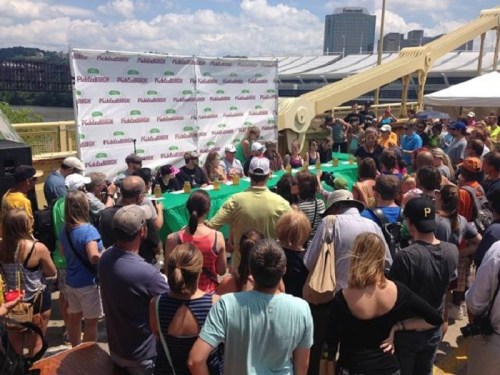 A large crowd gathers at an outdoor event with a panel of speakers behind a green table and a branded backdrop.