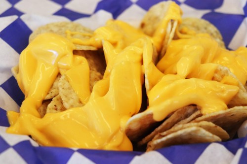 A basket of tortilla chips topped with melted cheese sauce on a blue and white checkered paper.