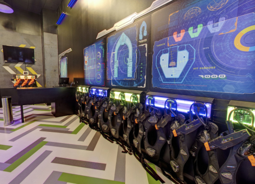 A gaming area featuring high-tech gear and screens, with vests hanging on a wall and a control station in the background.