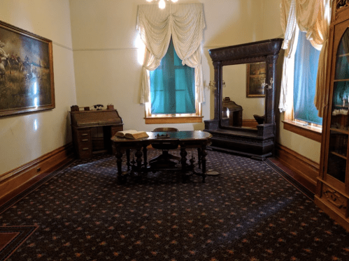 A vintage room with a wooden desk, mirror, and large window, featuring ornate curtains and a patterned carpet.