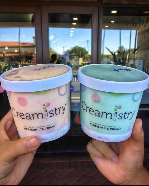 Two hands holding tubs of Creamistry premium ice cream, one light brown and one mint green, in front of a shop.