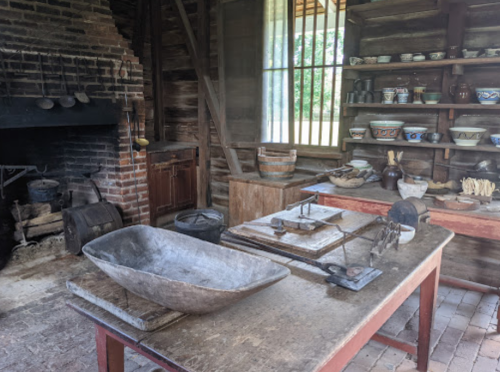 A rustic kitchen interior with a wooden table, fireplace, shelves of pottery, and various kitchen tools.