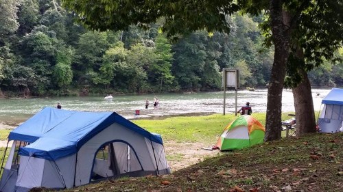 Tents set up by a riverbank, with people enjoying water activities and lush greenery in the background.