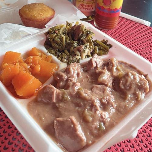 A takeout container with meat stew, collard greens, sweet potatoes, and a cornbread muffin on a red table.