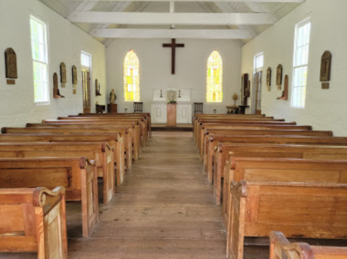 Interior of a simple church with wooden pews, a cross on the wall, and stained glass windows.