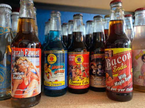 A collection of vintage soda bottles with colorful labels, including flavors like cream soda, cherry cola, and bacon soda.