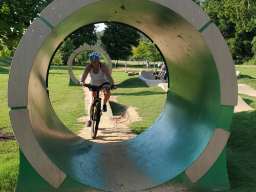 A cyclist rides through a large circular tunnel in a park, surrounded by green grass and trees.