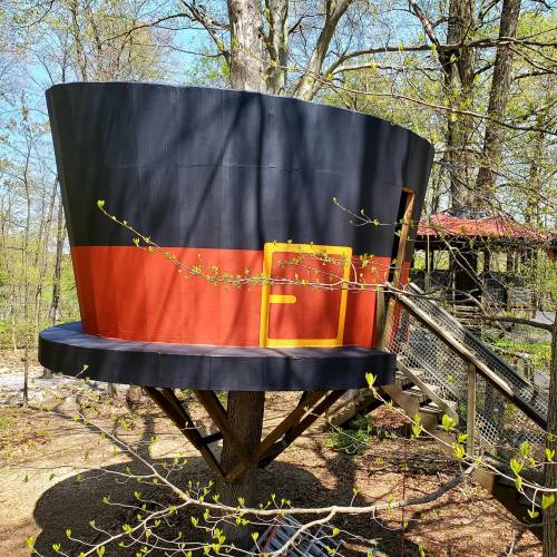 A whimsical treehouse resembling a large top hat, painted black and red, nestled among trees with a wooden walkway.