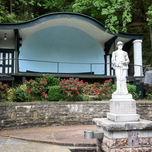 A stone statue of a soldier stands in front of a curved bandstand surrounded by colorful flowers and greenery.