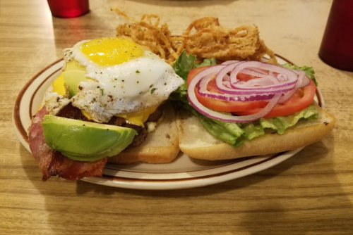 A sandwich with bacon, avocado, a fried egg, lettuce, tomato, and onion, served with crispy onion rings on the side.