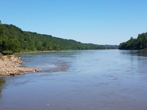 A calm river flows between lush green hills under a clear blue sky.