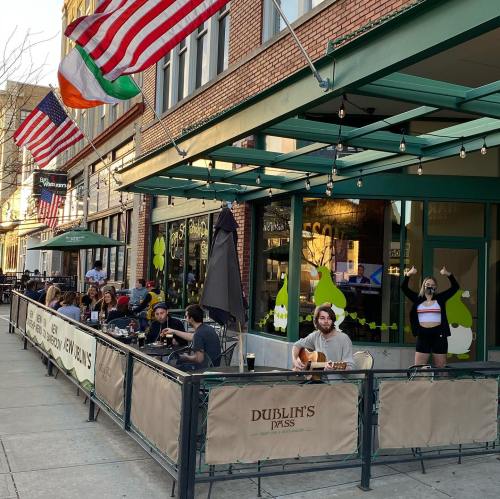 Outdoor dining scene at Dublin's Pub, with patrons enjoying food and drinks under string lights and flags.