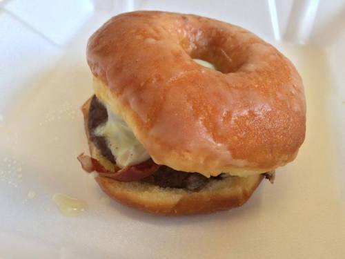 A glazed donut sandwich with a beef patty, bacon, and creamy sauce, served on a white takeout container.